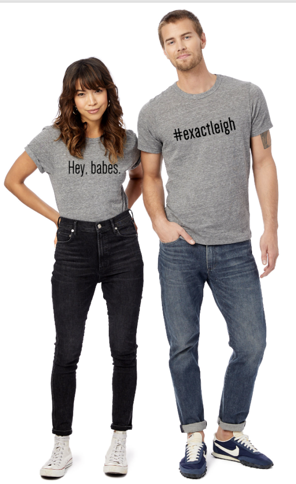 A brunette woman and a sandy haired man, both wearing jeans and grey t-shirts. Her shirt says, Hey, babes. His shirt says #exactleigh.
